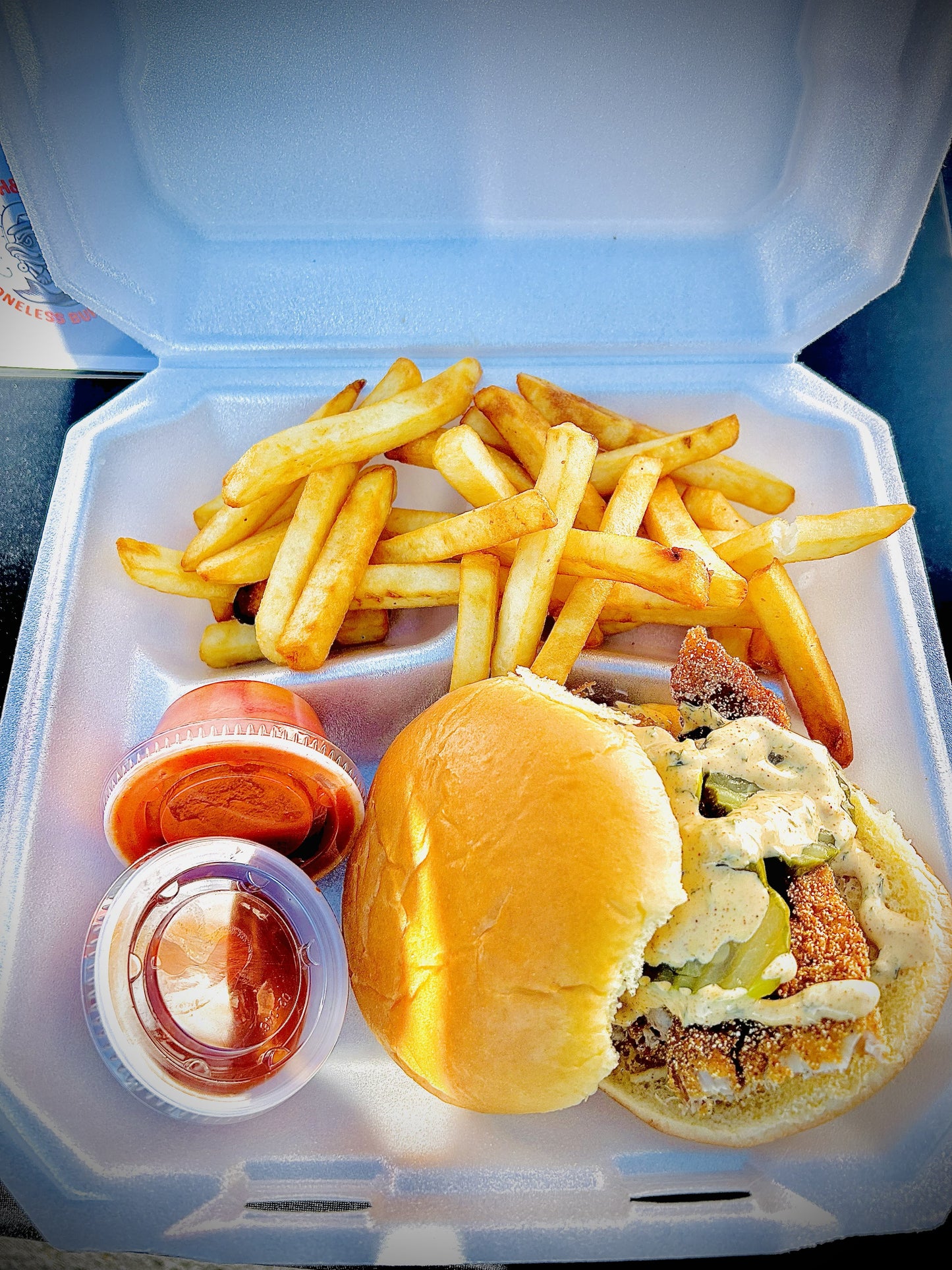 Buffalo Fish sandwich with fries, pickles and remoulade sauce in a Styrofoam container