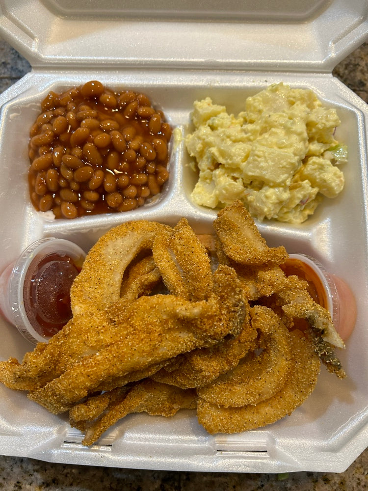 Take-out container with fried deboned boneless buffalo fish, baked beans, and potato salad on a stone surface.
