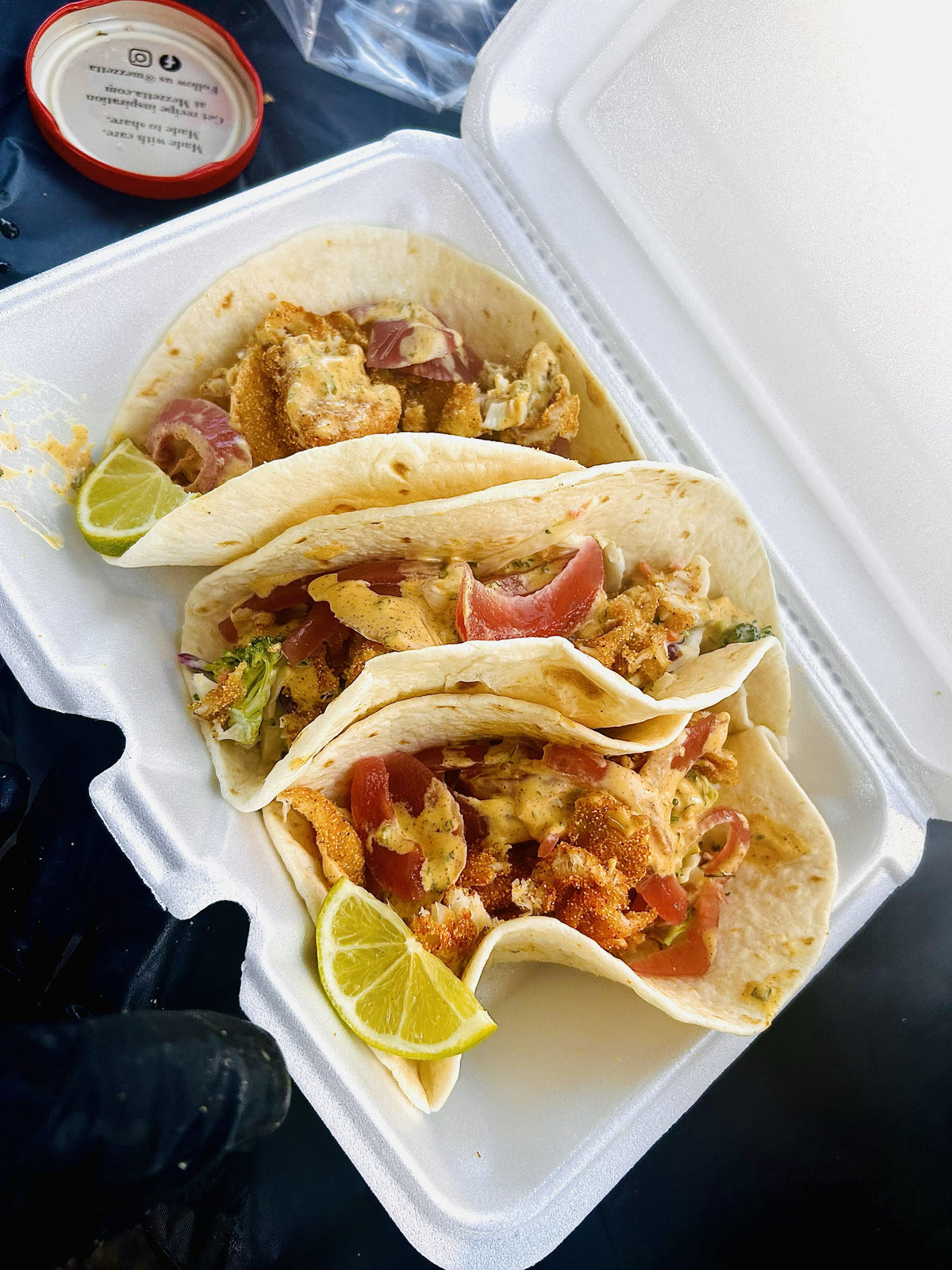Three buffalo fish tacos with Cole slaw, red pickled onions and taco sauce in a takeout container.